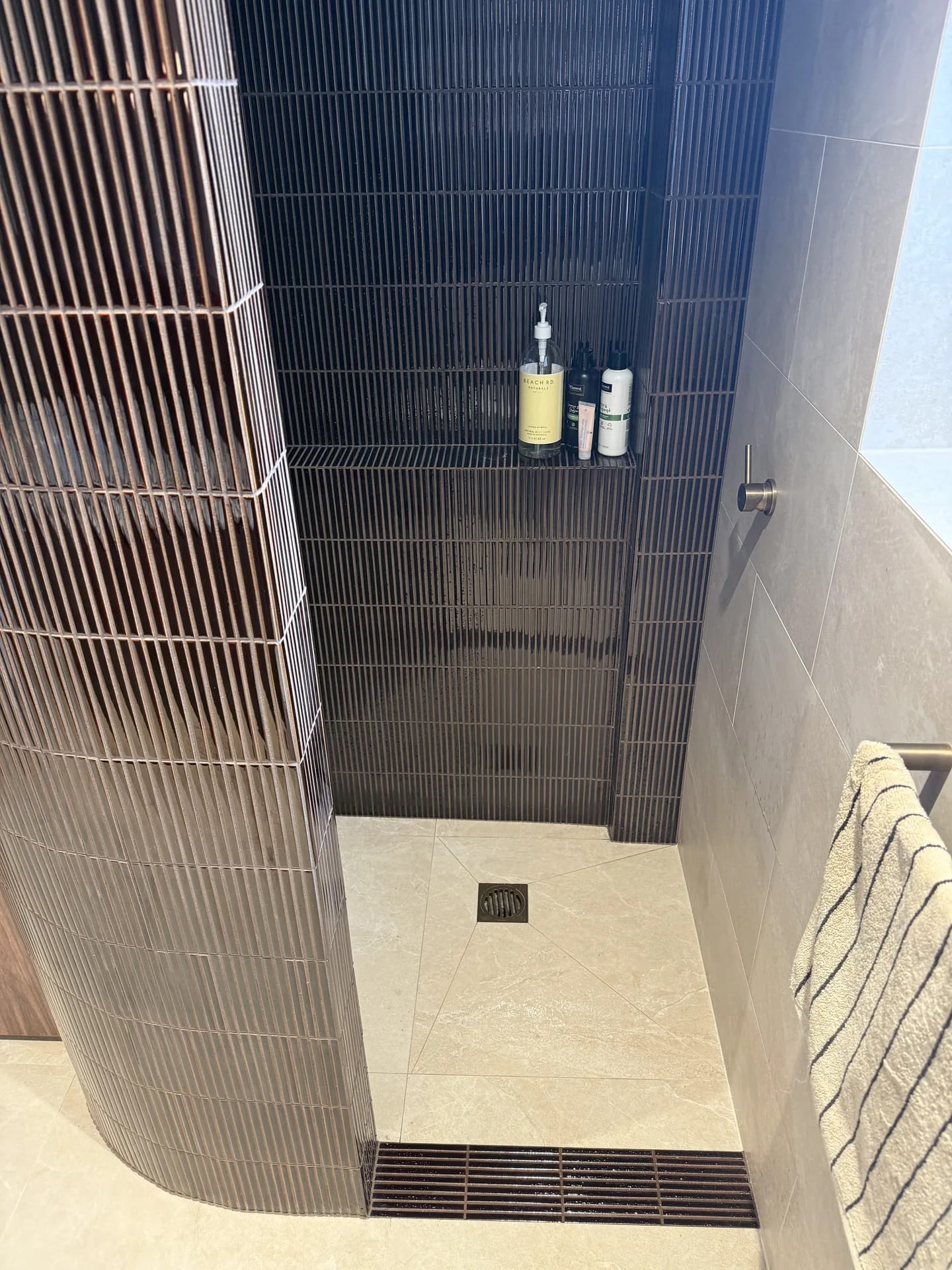 Walk-in shower entry with a curved dark finger-tiled wall, tiled shelf and linear floor grate