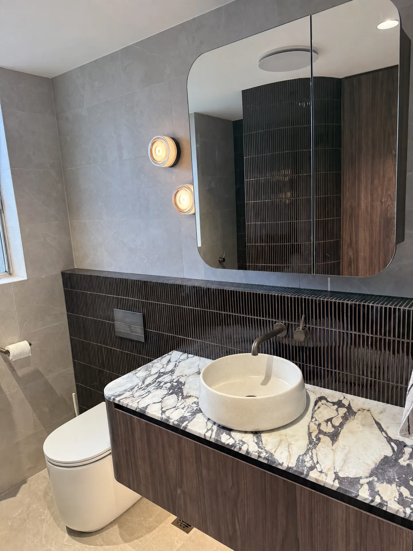 Vanity and wall-hung toilet against a dark finger tile band, with mirror cabinet and glass sconces