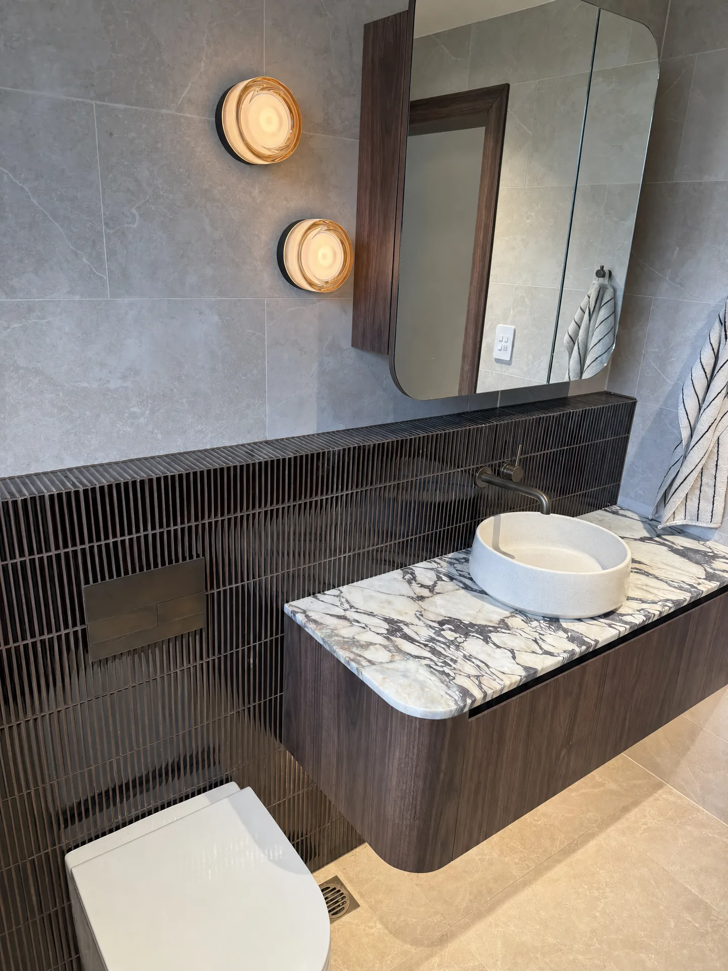 Wider view of the marble-topped vanity, wall-hung toilet and bronze flush plate on dark tile