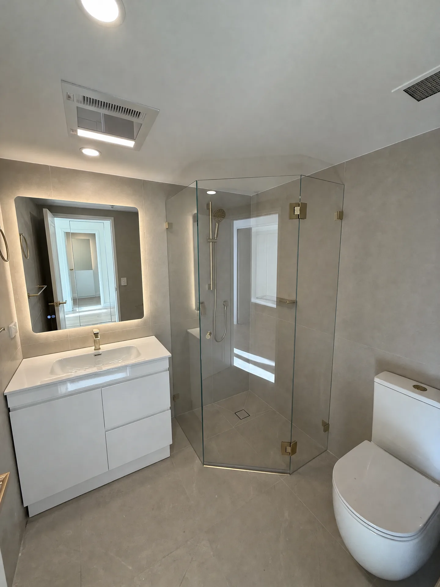 Bathroom with a backlit mirror, white vanity and an angled frameless corner shower