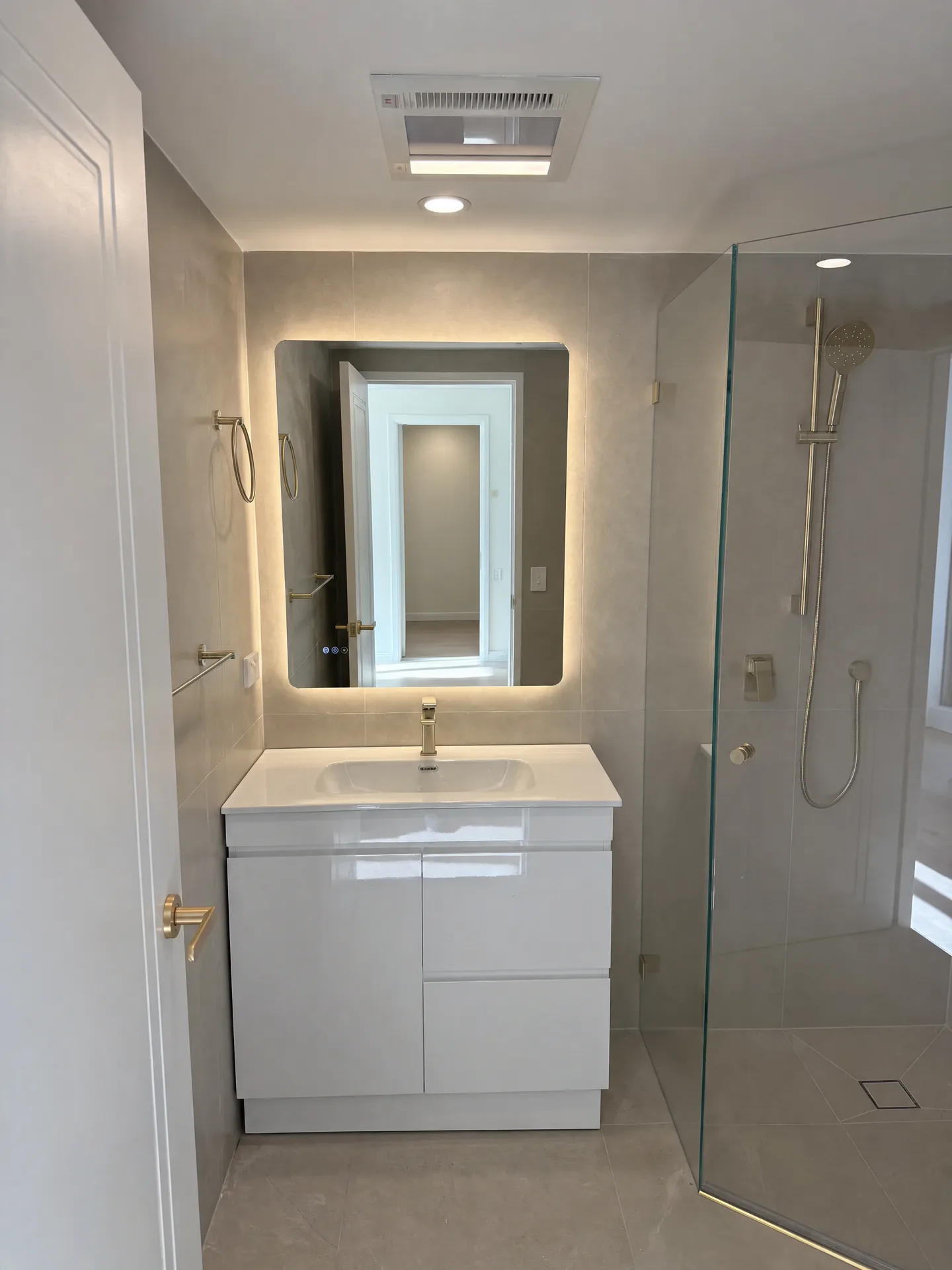 Vanity and backlit mirror beside a frameless glass shower with a brushed brass rail