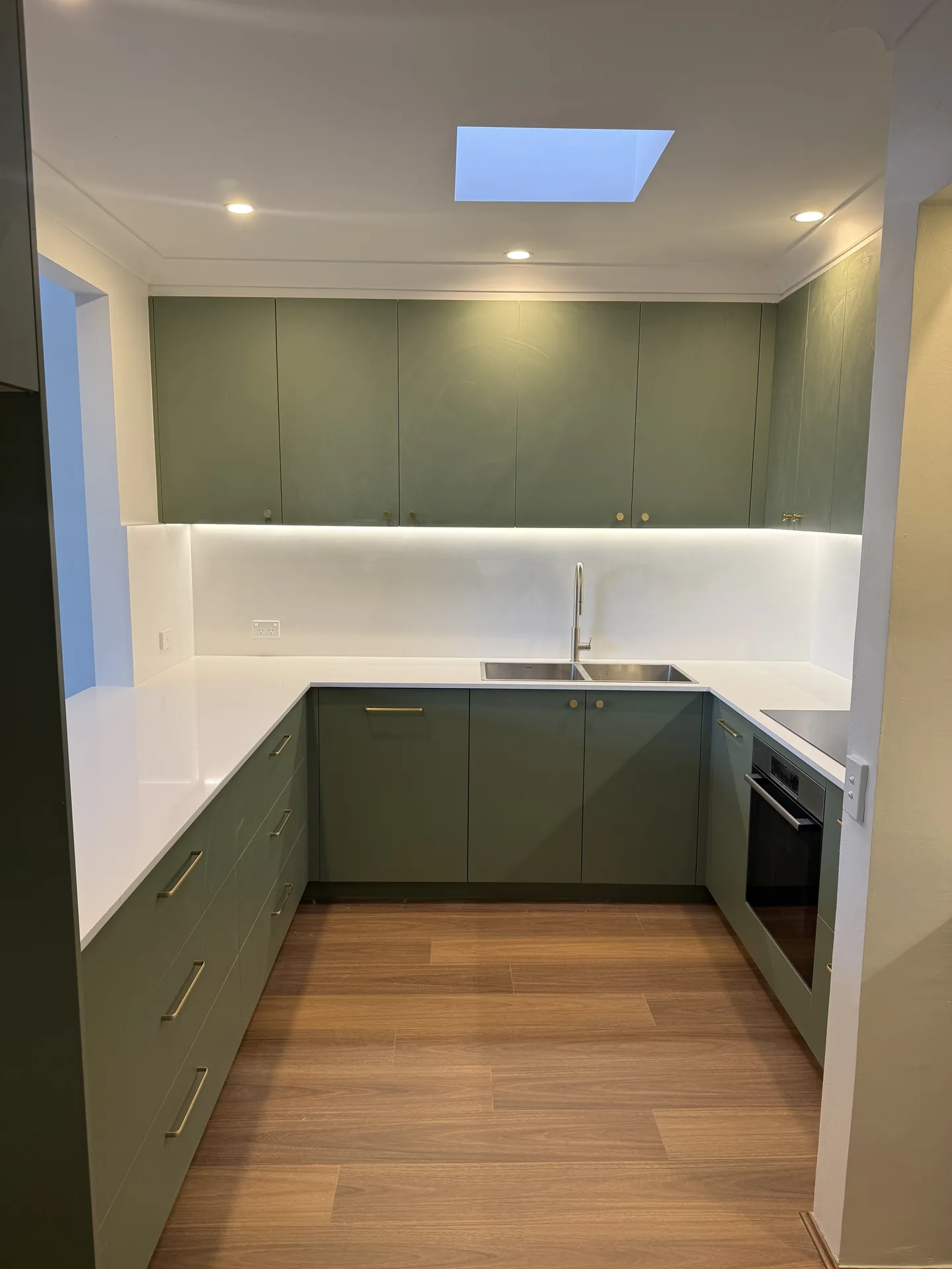 Olive green cabinetry on three walls of an apartment kitchen with brass knobs and a white benchtop