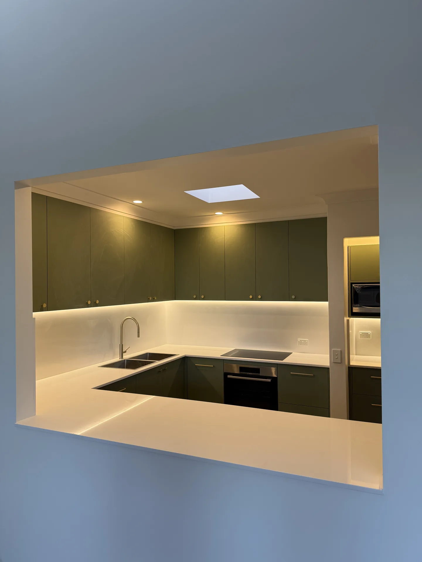 Kitchen seen through the servery opening, olive cabinetry and lit white splashback