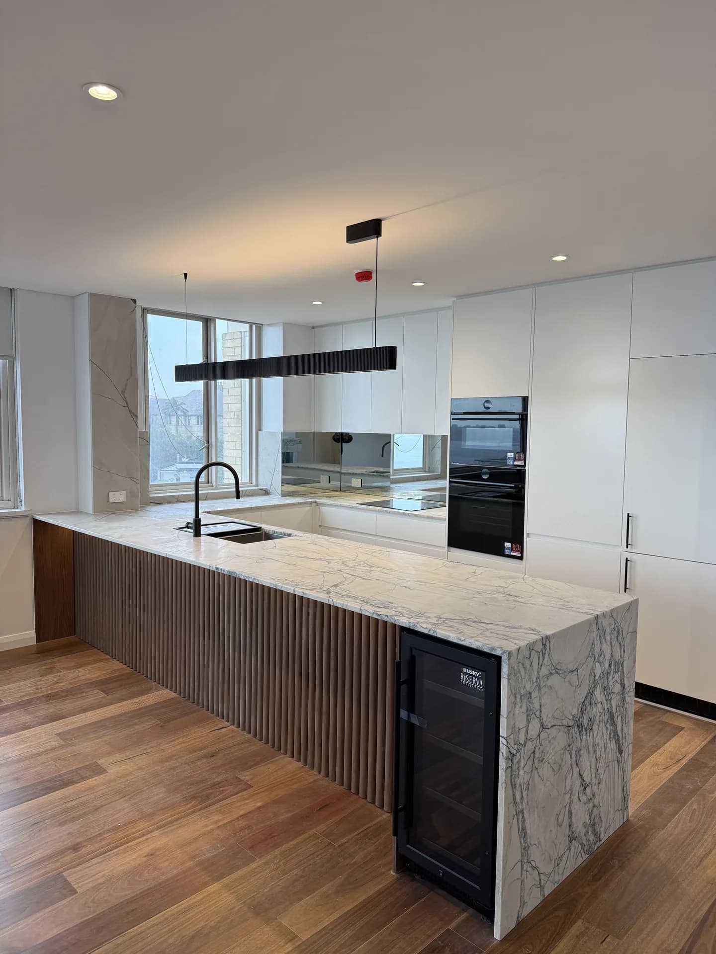 Kitchen island clad in fluted timber with a marble-look stone top and stacked black wall ovens behind