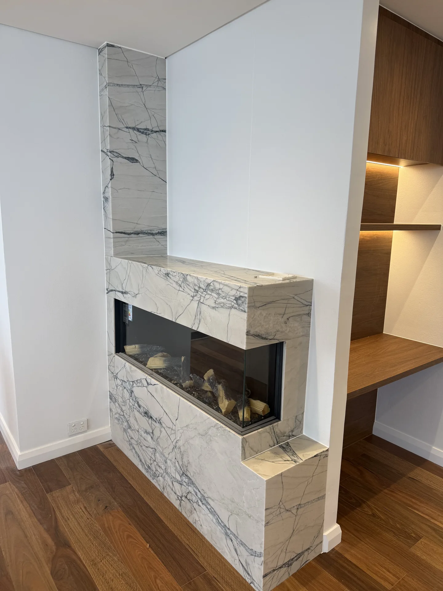 Fireplace clad in veined marble-look stone built into a dividing wall beside a timber desk nook