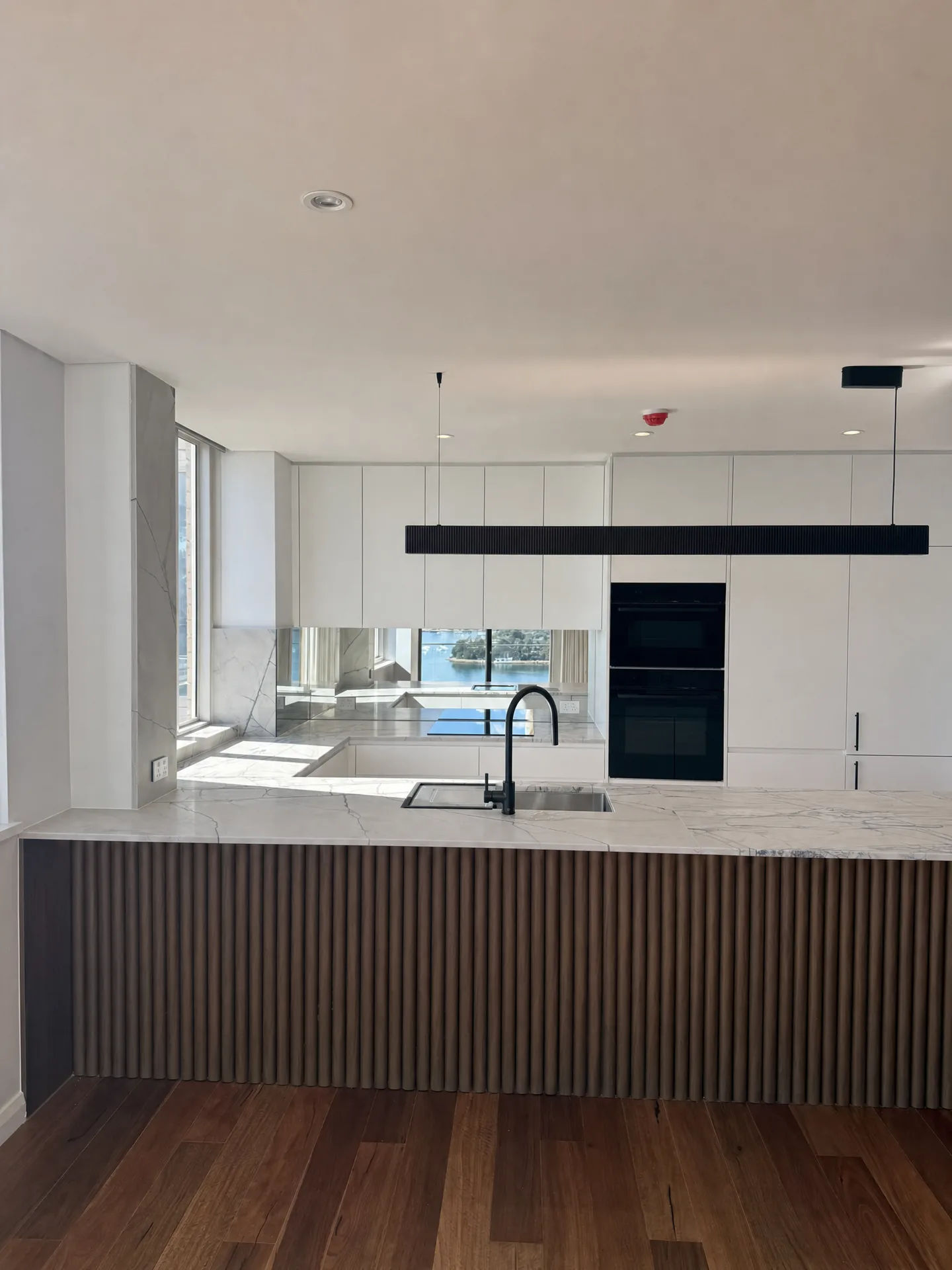 Black linear pendant over a stone-topped island with a mirrored splashback and harbour light behind
