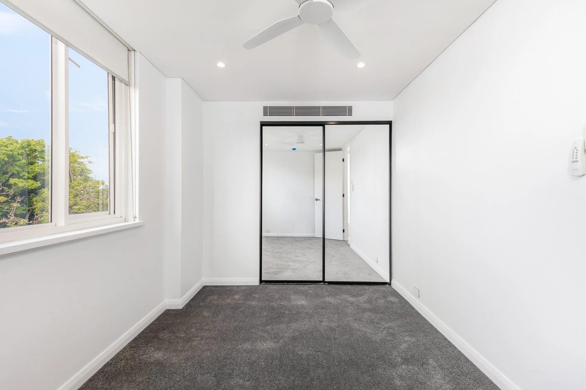 Empty bedroom with new charcoal carpet, a black-framed mirrored wardrobe and a ceiling fan