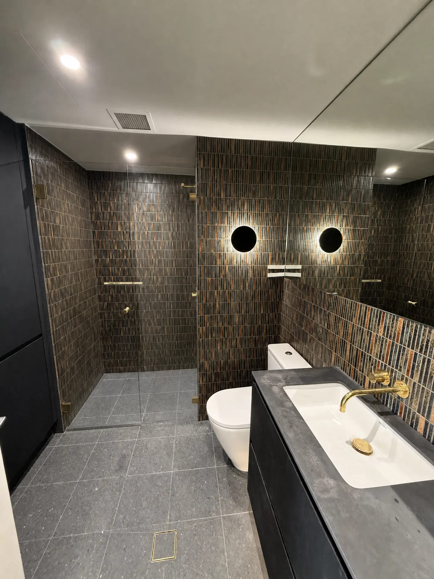 Frameless walk-in shower and wall-hung pan against bronze mosaic tiles with brass fittings