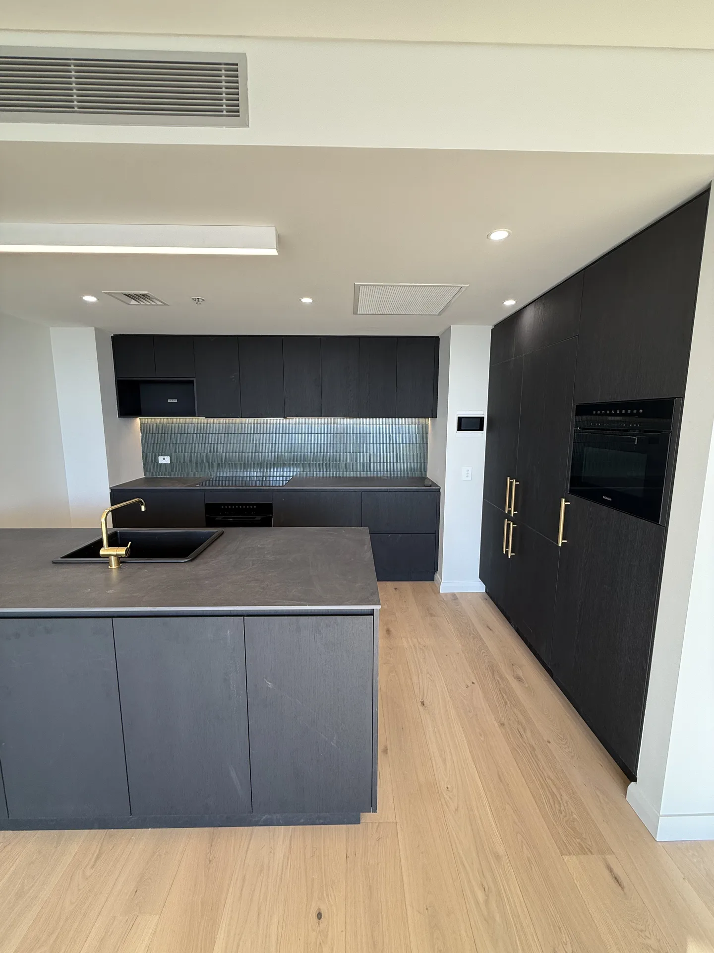 Charcoal timber-look kitchen joinery with brass handles, integrated oven and green splashback