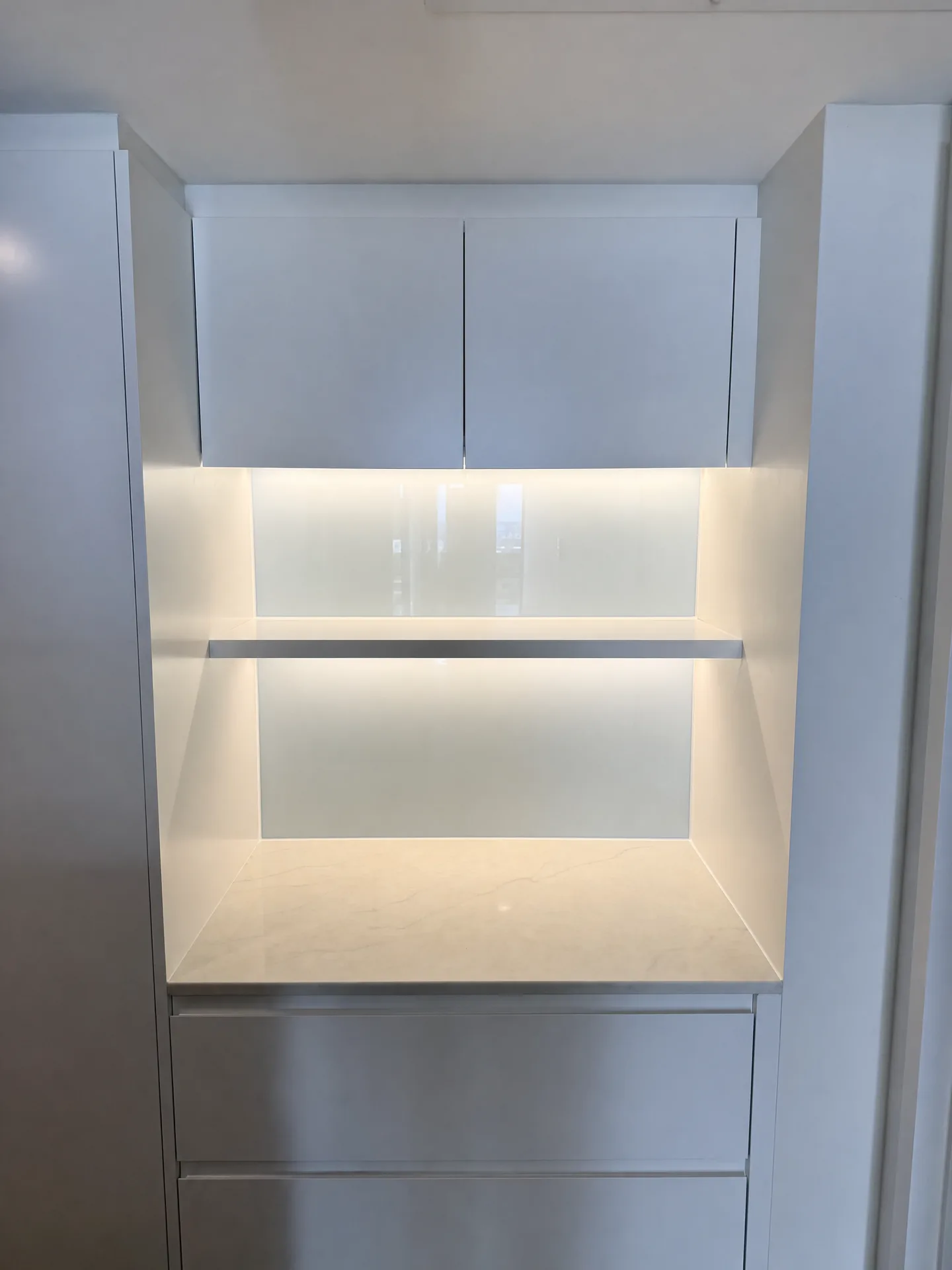 Lit bench-height niche with glass back, slim shelf and stone shelf set into white cabinets