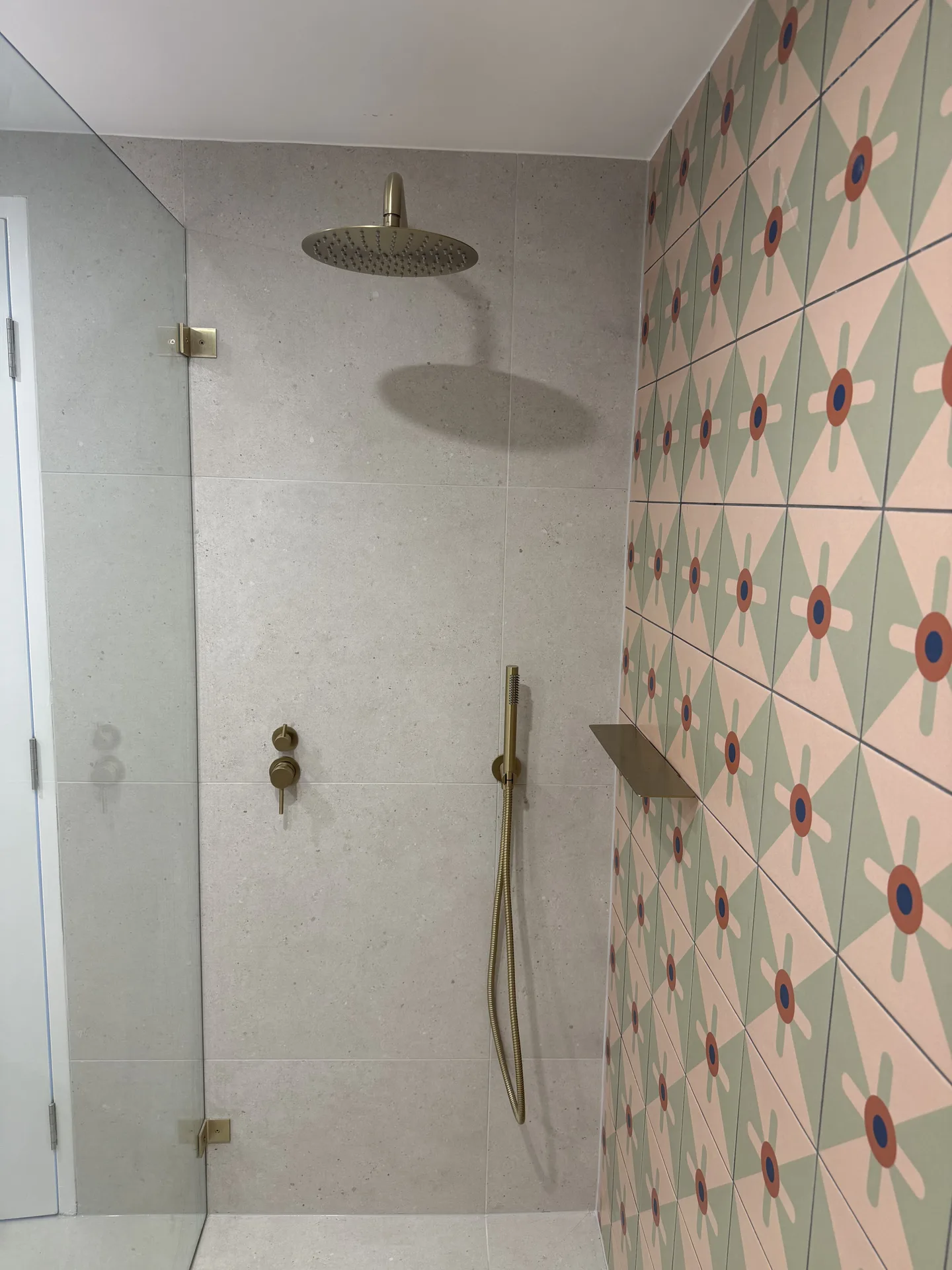 Walk-in shower with brushed brass rain head beside a green and pink patterned feature wall