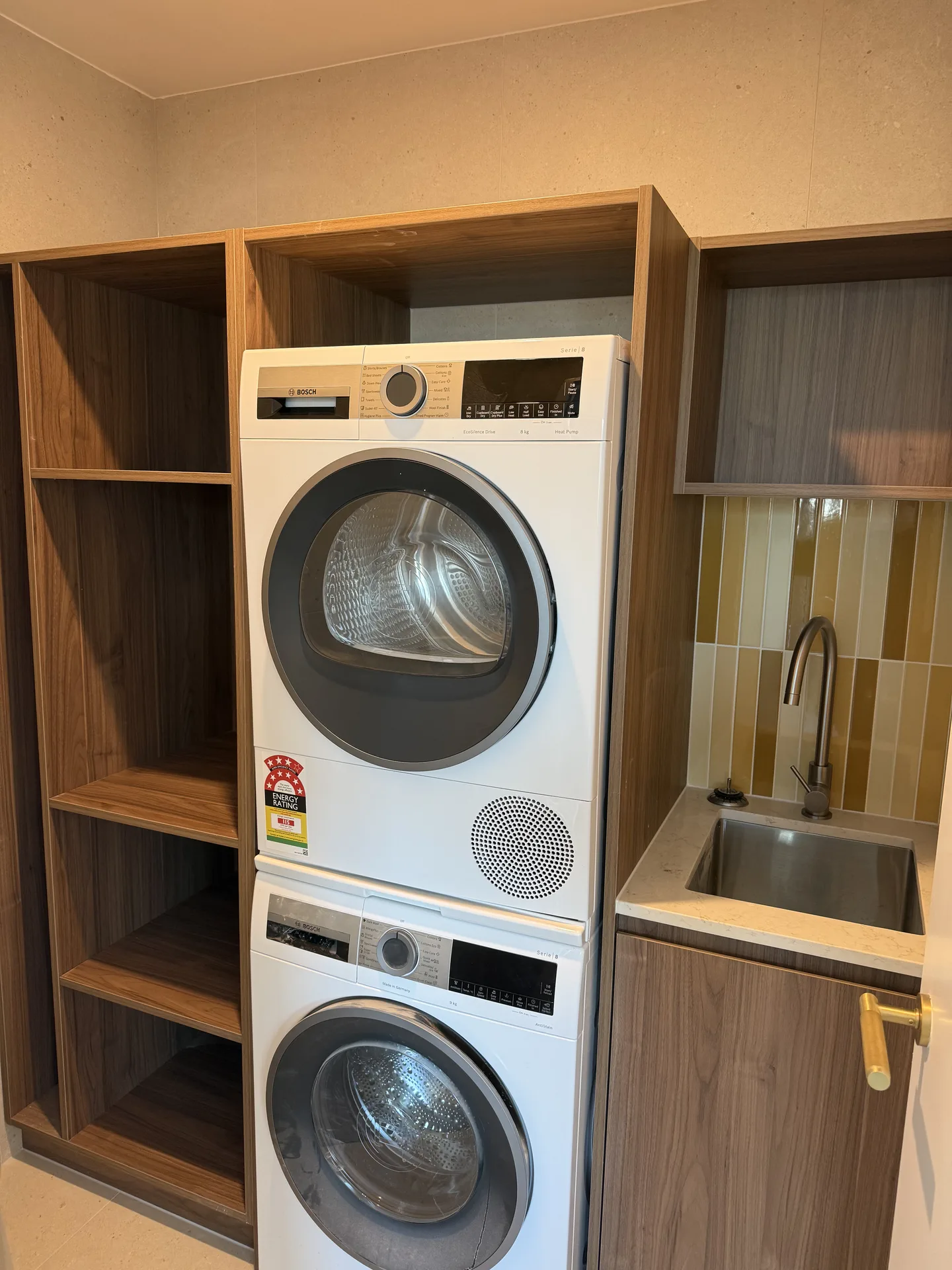 Stacked washer and dryer in timber-look joinery beside a sink with ochre vertical tiles